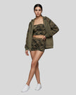 A woman modeling a matching Athlesi camouflage activewear set consisting of a tube top, shorts, and an open snap-button hoodie, paired with white sneakers, against a solid gray studio background.