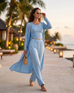 Woman in a light blue outfit walking along a tropical path with palm trees and lanterns in the background.