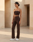 A woman stands in a modern architectural setting with beige stone walls, wearing a matching brown Contour Yoga Tube Top and Contour Yoga Tube Pants, paired with white sneakers.