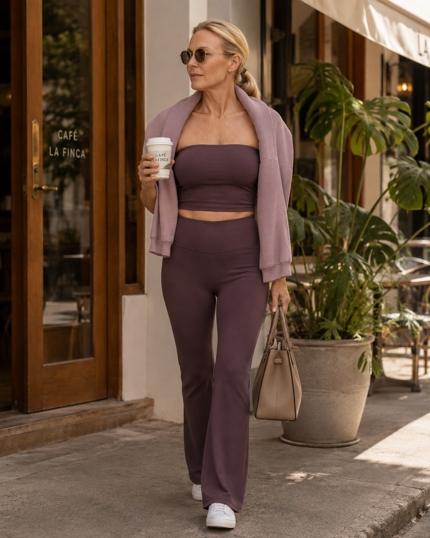 A woman walking on a city sidewalk wearing a matching plum purple Contour Yoga set consisting of a strapless tube top and high-waisted yoga pants, with a light mauve cardigan draped over her shoulders, white sneakers, and a beige handbag.