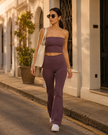 A woman walking on a sunlit street wearing a matching plum purple Contour Yoga, consisting of a strapless tube top and high-waisted flared yoga pants set, carrying a cream shoulder bag.