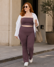 A woman walking on a city sidewalk wearing a matching plum purple Contour Yoga set consisting of a strapless crop top and high-waisted yoga pants, styled with an open white shirt, white sneakers, sunglasses, and a beige tote bag.