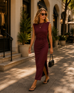 A woman walks on a sunlit city sidewalk wearing a Wine Red ContourEase Ruched Maxi Dress. She is styled with black sunglasses, layered gold necklaces, black sandals, and a small black handbag.