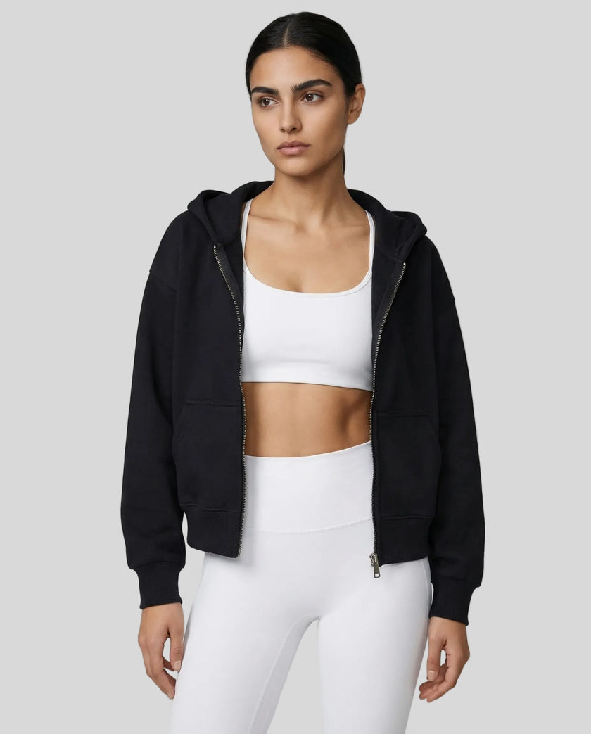 Model wearing a black CoreZip Hooded Jacket over a white sports bra and leggings on a neutral setting.