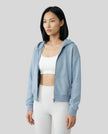 Model wearing a blue CoreZip Hooded Jacket over a white sports bra and leggings on a neutral setting.