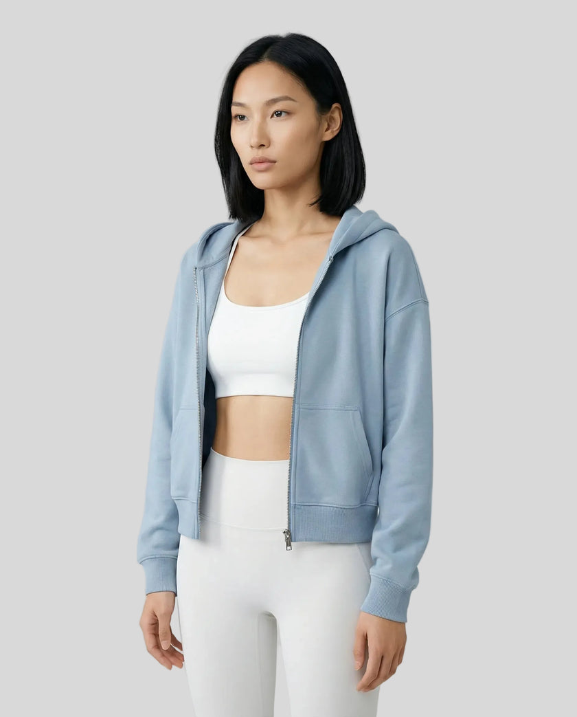 Model wearing a blue CoreZip Hooded Jacket over a white sports bra and leggings on a neutral setting.