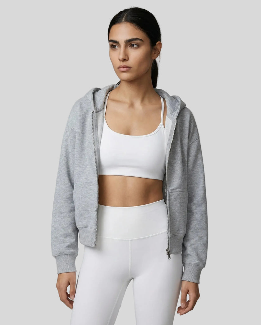 Model wearing a gray CoreZip Hooded Jacket over a white sports bra and leggings on a neutral setting.