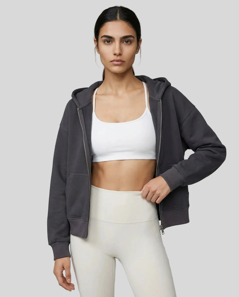 Model wearing a dark gray CoreZip Hooded Jacket over a white sports bra and leggings on a neutral setting.