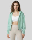 Model wearing a light green CoreZip Hooded Jacket over a white sports bra and leggings on a neutral setting.