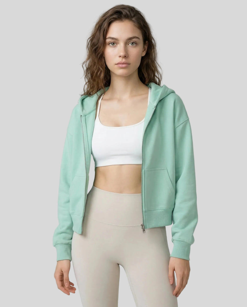Model wearing a light green CoreZip Hooded Jacket over a white sports bra and leggings on a neutral setting.