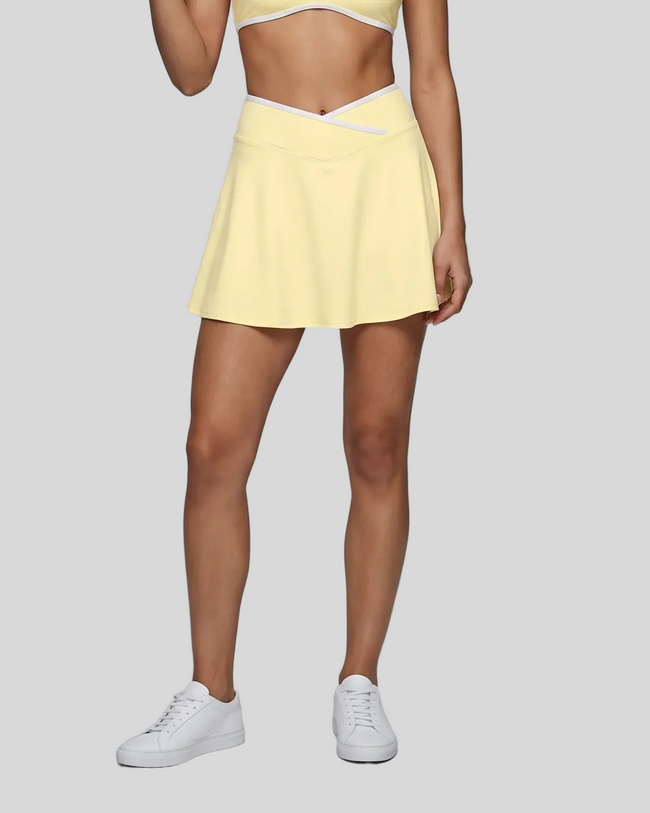 Lower body view of a model wearing a Light Yellow CourtClub Color Block A-Line Skirt, paired with a matching Light Yellow CourtClub Color Block Sports Bra and white sneakers.