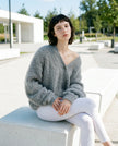 Woman wearing a dark gray sweater and white pants sitting on a bench outdoors.