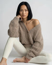 Woman wearing a khaki cardigan and white pants sitting on a white background