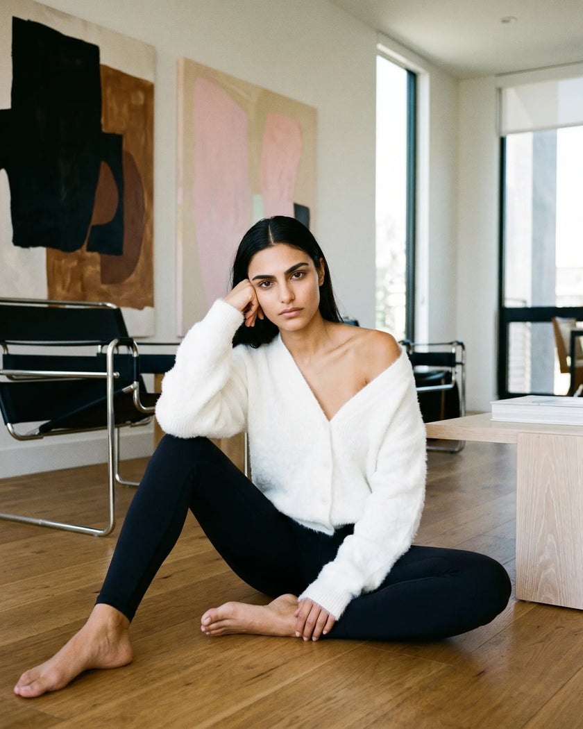 Woman wearing EmberSoft Cardigan in white, sitting on a wooden floor in a modern living room with abstract art on the wall.