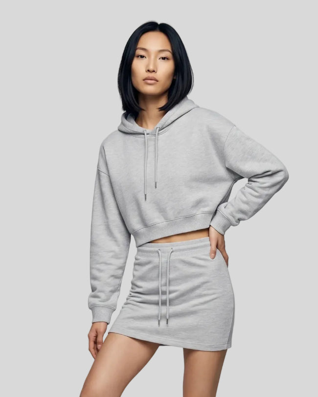 A woman modeling a matching flower gray two-piece loungewear set consisting of Everyday Cropped Hoodie and Everyday Drawstring Skirt, paired with white sneakers.