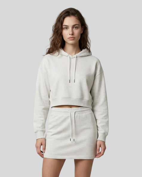 A woman modeling a matching oat gray two-piece loungewear set consisting of Everyday Cropped Hoodie and Everyday Drawstring Skirt, paired with white sneakers.