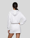 A model from behind wearing a white Everyday Cropped Hoodie and matching Everyday Drawstring mini skirt set, paired with white sneakers.