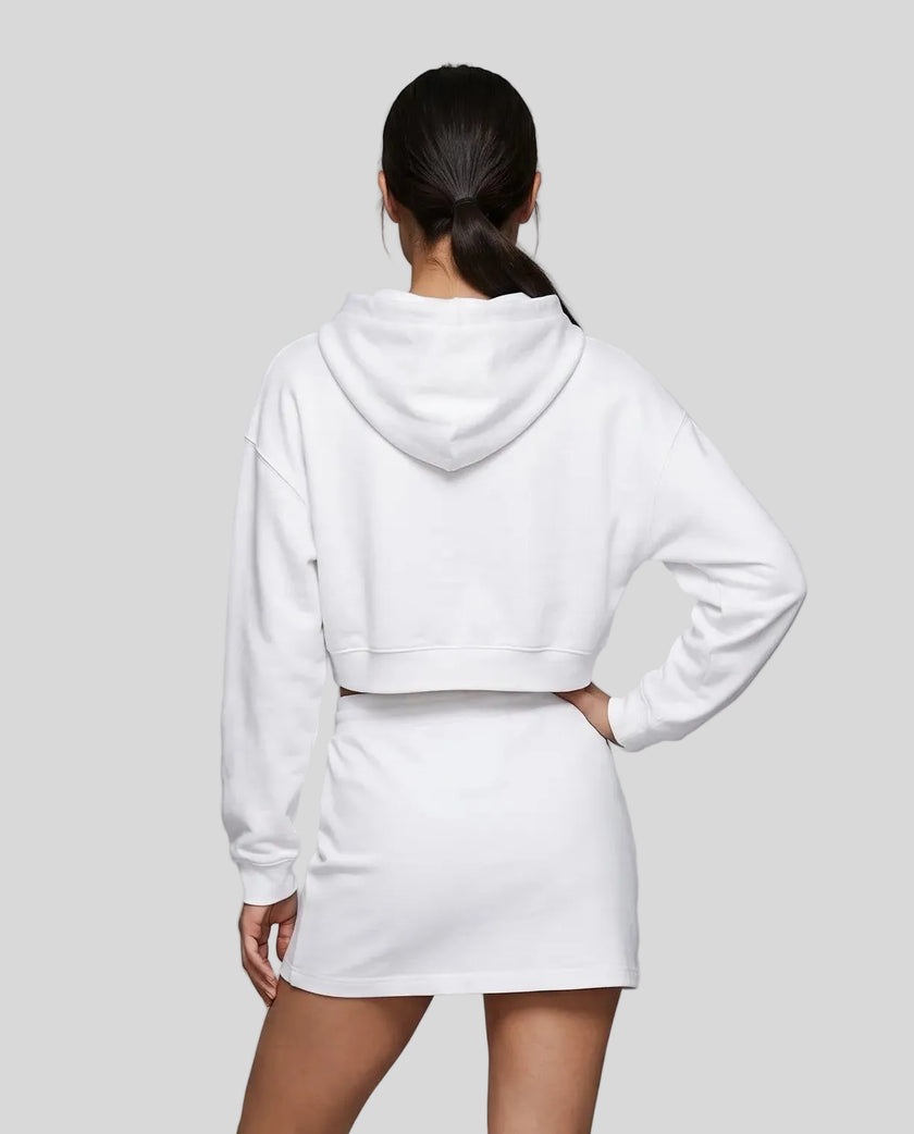 A model from behind wearing a white Everyday Cropped Hoodie and matching Everyday Drawstring mini skirt set, paired with white sneakers.