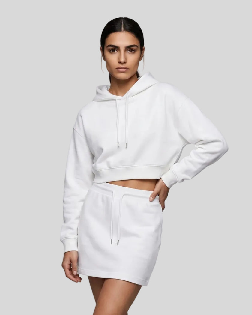 A woman modeling a matching white two-piece loungewear set consisting of Everyday Cropped Hoodie and Everyday Drawstring Skirt, paired with white sneakers.