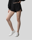 A model wearing black athletic shorts with a drawstring waist, paired with a black cropped sweatshirt and white sneakers, shown from the waist down against a solid light gray background.