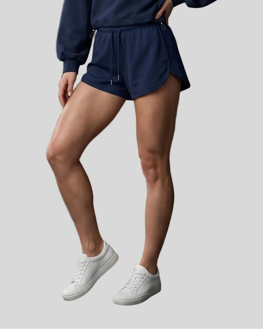 A model wearing navy blue athletic shorts with a drawstring waist, paired with a matching navy sweatshirt and white sneakers, shown from the waist down against a solid light gray background.