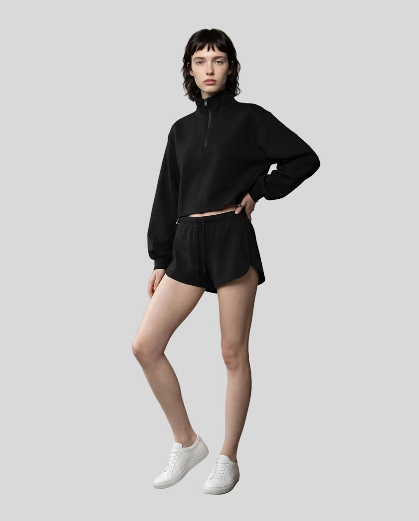Full body view of a woman modeling a black Athlesi Heritage Quarter-Zip Sweatshirt and matching shorts set, paired with white sneakers.