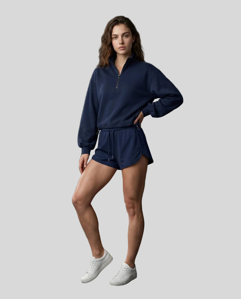 Full body view of a woman modeling a navy blue Athlesi Heritage Quarter-Zip Sweatshirt and matching shorts set, paired with white sneakers.