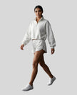 Full body view of a woman modeling a white Athlesi Heritage Quarter-Zip Sweatshirt and matching shorts set, paired with white sneakers.