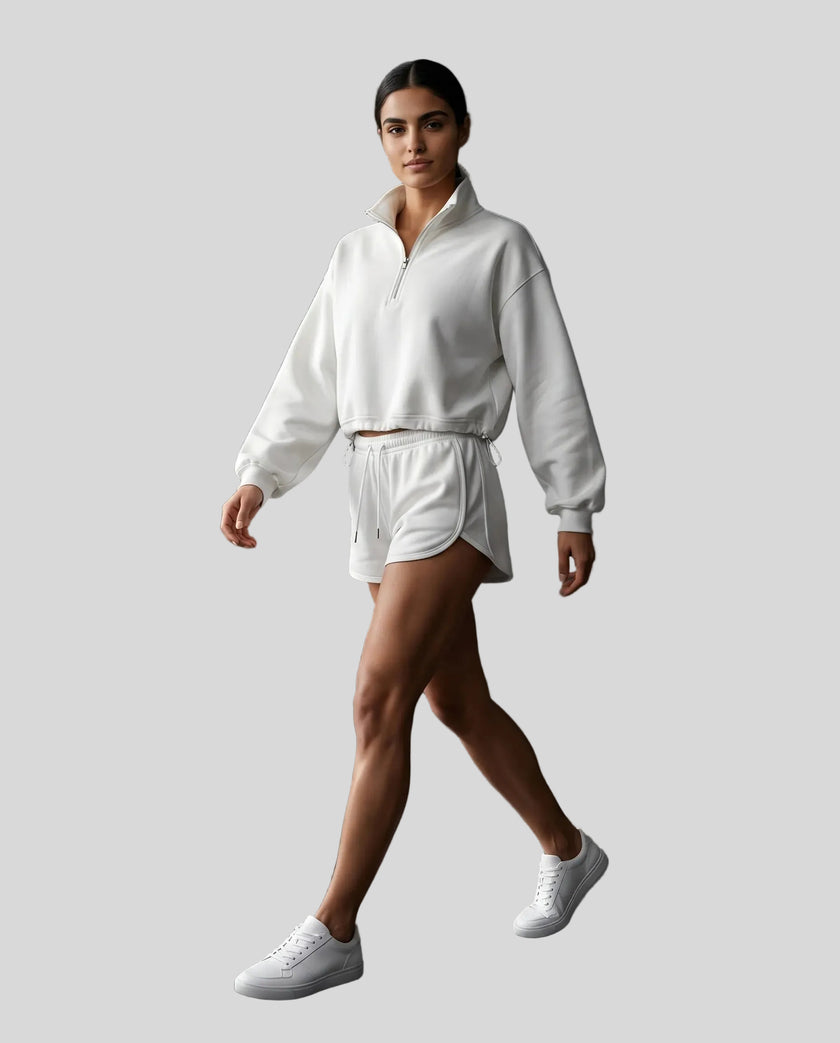Full body view of a woman modeling a white Athlesi Heritage Quarter-Zip Sweatshirt and matching shorts set, paired with white sneakers.