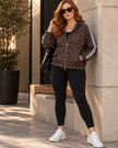 A woman stands outdoors against a beige stone wall wearing Athlesi Leopard Accent Hoodie in leopard brown with white stripes on the sleeves, black leggings, and white sneakers.