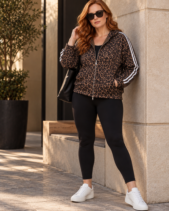 A woman stands outdoors against a beige stone wall wearing Athlesi Leopard Accent Hoodie in leopard brown with white stripes on the sleeves, black leggings, and white sneakers.
