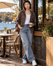 A woman stands on a lakeside patio wearing a Athlesi Leopard Accent Hoodie in Light Taupe over a white tank top, light-wash wide-leg jeans, and white sneakers, while holding a coffee cup.