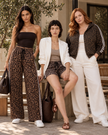 Three women modeling coordinated outfits in an outdoor patio setting. The outfits feature Athlesi Leopard Accent pieces, including barrel pants, shorts, and a zip-up hoodie with white stripes, paired with solid white or neutral tops.