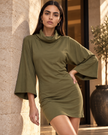 A woman models an army green Muse Cowl Neck Tunic dress with wide bell sleeves, posing against a beige stone wall in a warm, sunlit setting.