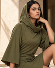 A woman wearing an army green hooded Muse Cowl Neck Tunic dress with wide bell sleeves, posing in a warm, sunlit architectural setting.