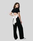 Angled full body back view of woman wearing Nova Tie-Back Tee in Black, showing the open-back silhouette and White tie