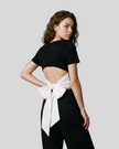 Angled upper body back view of woman wearing Nova Tie-Back Tee in Black, showing the open-back silhouette and White tie