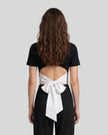Upper body back view of woman wearing Nova Tie-Back Tee in black, with open-back cutout and contrasting White bow