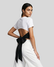 Angled upper body back view of woman wearing Nova Tie-Back Tee in white, showing the open-back silhouette and Black tie