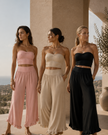 Three women model matching QuietForm Ruched two-piece outfits on a sunlit terrace with a Mediterranean-style landscape in the background. The outfits are wide-leg cropped pants with tube tops.