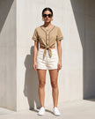 A woman stands outdoors against a concrete wall wearing a Coffee TrackLine Baseball Jersey, paired with white shorts and white sneakers.