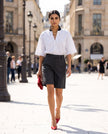 A woman walking down a sunny city street wearing a white TrackLine Baseball Striped Jersey, black high-waisted shorts, and red pointed-toe heels, holding a red clutch.