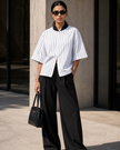 A woman stands outdoors wearing a TrackLine Baseball Striped Jersey, black wide-leg pants, white sneakers, and a black handbag.