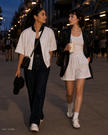 Two women walking together in an outdoor stadium or event area at dusk, wearing coordinated streetwear. One wears a TrackLine Baseball Striped Jersey over a black top with dark jeans, and the other wears a TrackLine Baseball Striped Shorts with a white top and a black leather jacket.