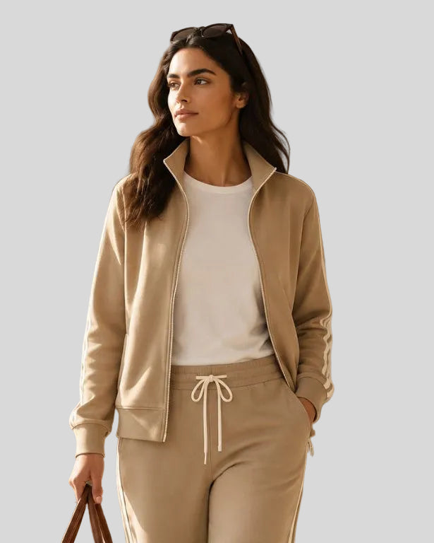 A woman in a beige Athlesi TrackLine Club Jacket Sweatpants, walks through a sunlit indoor space while carrying a brown leather duffel bag. 