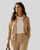 A woman in a beige Athlesi TrackLine Club Jacket Sweatpants, walks through a sunlit indoor space while carrying a brown leather duffel bag. 