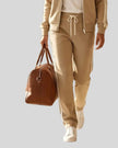 A woman walking wearing a khaki TrackLine Club sweatpants over a white top, carrying a brown leather bag.