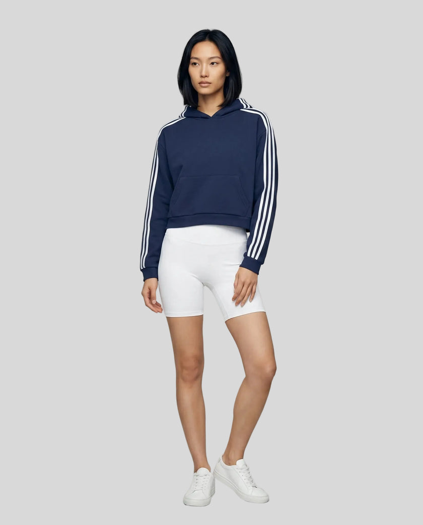 Full body front view of a woman wearing the Athlesi TrackLine Cropped Hoodie in blue styled in a modern architectural setting
