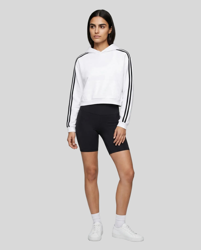 Full body front view of a woman wearing the Athlesi TrackLine Cropped Hoodie in white styled in a modern architectural setting