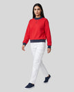 Woman modeling a red TrackLine Heritage Sweatshirt with navy blue ribbed trim, paired with white pants and navy-blue sneakers.